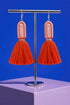 A pair of vibrant orange tassel earrings with a metallic frame, displayed on a stand against a solid blue background.