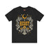 A unisex short sleeve graphic t shirt in dark charcoal black featuring a highly detailed symmetrical medallion print on the chest. In the center, the word RESIST is printed in a bold yellow gothic blackletter font. Two coiled white snakes with fine black scale details frame the text on the left and right. Above the text, a yellow crescent moon is flanked by white and yellow starbursts. Beneath, three yellow marigold flowers sit atop white stems and leafy foliage.