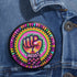 Round pinback button with psychedelic rainbow design featuring a raised black fist surrounded by concentric circles in vibrant pink, orange, green, and yellow. The word "RESIST" appears in white text below the fist, set against black background with flower details. Activist protest pin with glossy finish and safety pin backing.