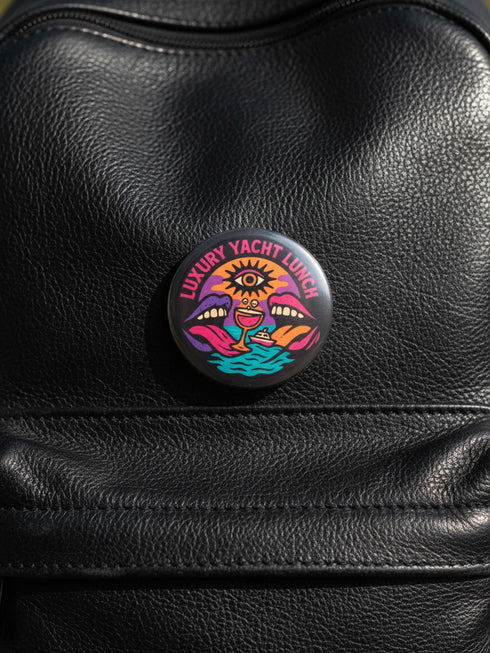 A round 1.75 inch glossy pinback button featuring satirical psychedelic artwork on a black background. The text LUXURY YACHT LUNCH arches over the top in bold neon pink letters. The central design depicts a surreal scene with a stylized third-eye sun, large open mouths with tongues sticking out, a smiling cocktail glass character, and a small white yacht floating on wavy teal water. The color palette includes retro 70s neon pink, orange, purple, and teal. The button has a safety pin backing.