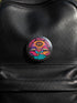 A round 1.75 inch glossy pinback button featuring satirical psychedelic artwork on a black background. The text LUXURY YACHT LUNCH arches over the top in bold neon pink letters. The central design depicts a surreal scene with a stylized third-eye sun, large open mouths with tongues sticking out, a smiling cocktail glass character, and a small white yacht floating on wavy teal water. The color palette includes retro 70s neon pink, orange, purple, and teal. The button has a safety pin backing.