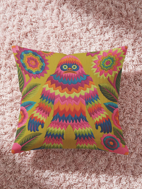 A square faux embroidery pillow featuring a brightly colored maximalist design against a mustard yellow background. In the center stands a stylized, furry Bigfoot or Sasquatch figure rendered in vibrant layers of pink, orange, blue, and lime green with a blue face and yellow eyes. The central creature is surrounded by large, detailed symmetrical pink and blue flowers and leaves. The flat printed fabric creates a highly realistic optical illusion of dimensional crewel embroidery stitches.