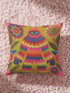 A square faux embroidery pillow featuring a brightly colored maximalist design against a mustard yellow background. In the center stands a stylized, furry Bigfoot or Sasquatch figure rendered in vibrant layers of pink, orange, blue, and lime green with a blue face and yellow eyes. The central creature is surrounded by large, detailed symmetrical pink and blue flowers and leaves. The flat printed fabric creates a highly realistic optical illusion of dimensional crewel embroidery stitches.