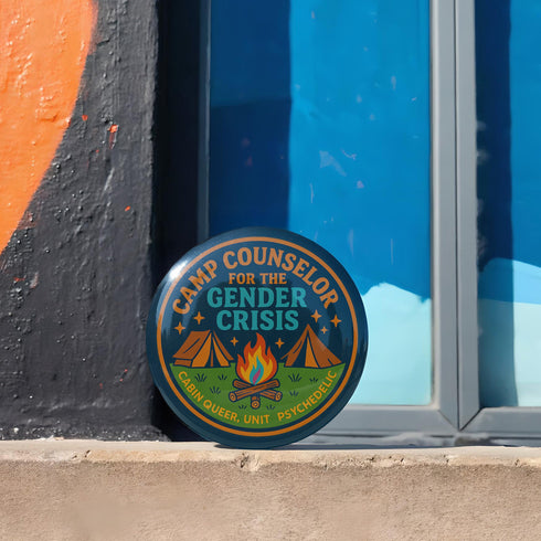 Round glossy pinback button measuring 2.25 inches featuring a playful camp-themed design with the phrase “Camp Counselor for the Gender Crisis.” The artwork includes two orange tents, a rainbow campfire, stars, and green grass. Bottom text reads “Cabin Queer, Unit Psychedelic.” Made from durable tinplate with a safety pin back and coated in a UV and scratch-resistant mylar finish. Bold and colorful, perfect for jackets, bags, or pride outfits.