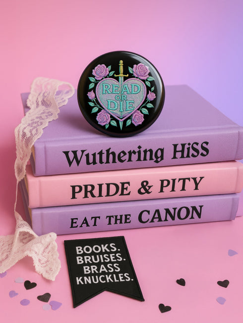 Glossy black pinback button featuring embroidered-style artwork of a pastel purple heart that reads “Read or Die” pierced by a sword and surrounded by roses; shown on books, graffiti, and lace backgrounds.