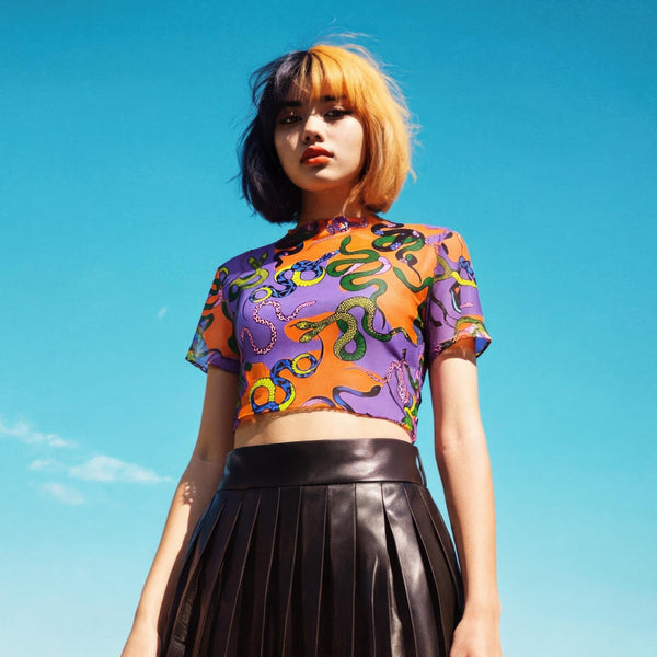 A vibrant see-through mesh crop top with a high mock neck and short sleeves. The shirt features a bold, all-over snake print pattern with colorful green, blue, yellow, and black serpents on a striking orange and purple color-block background. This Y2K-style top is a statement piece of witchy, goth, and alternative clothing, perfect for a maximalist rave outfit or edgy festival fashion from a black owned shop.