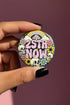 A round pinback button featuring a solid black background covered in a retro pastel colored design. The center reads 25TH NOW in a groovy bubbly font using pale pink and mint green referencing the 25th amendment. Surrounding the text is a maximalist cluster of cute but edgy motifs including smiling white skulls, pastel rainbows, pale yellow crescent moons, daisies, and a pink and white spotted mushroom. The button has a glossy laminate finish and a secure metal safety pin backing.