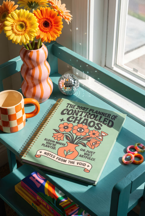 A sage green hardcover spiral planner for 2027-2028 with the title "The 2027 Planner of Controlled Chaos" in a funky, retro 70s-style font. The cover features a groovy illustration of orange and pink flowers in a vase, with one flower depicted as an eyeball. The text reads "Bloom where you're planted. Or just wilt artfully." and "Notes from the Void." The planner has a gold metal wire-o binding. The back cover shows a decorative floral frame with a "Property Of" section