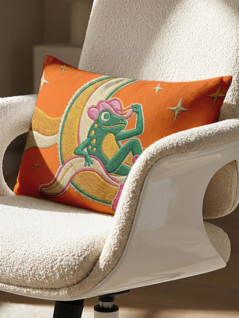 Rectangular 20x12 inch lumbar pillow cover in bright orange featuring printed faux embroidery of a green frog wearing a pink cowgirl hat and pink cowboy boots, lounging on a golden yellow crescent moon shaped like a peeled banana, surrounded by cream and gold sparkle stars and retro wavy stripes. Whimsical cowboy frog design printed on both sides with a hidden zipper, crafted from soft linen look polyester in a funky 70s retro style.