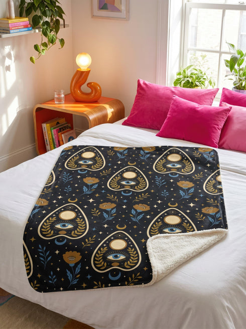 A rectangular velveteen minky blanket featuring a repeating dark academia pattern on a deep black background. The design includes gold and cream Ouija planchettes, each containing a blue all-seeing eye, a cream circle, and a light blue crescent moon framed by botanical leaves. Scattered between the planchettes are gold flowers with light blue stems, additional crescent moons, and small four-point stars. The reverse side of the blanket is made of a fluffy, textured off-white sherpa fleece.