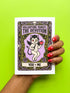 A vertical folded greeting card featuring a pastel goth tarot design titled The Devotion. The central illustration shows a pale devil imp with black wings and horns, holding a red heart in the left hand and a black dagger in the right. The imp has one star eye and one heart eye. The background is lavender purple. A black border frames the scene with teal vines, skulls, gold crescent moons, and roses. The bottom banner reads YOU + ME. The finish is shimmering pearlescent.