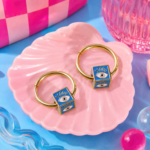 two gold hoop earrings with blue eye-shaped charms, placed on a pink shell-shaped plate.