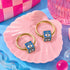 two gold hoop earrings with blue eye-shaped charms, placed on a pink shell-shaped plate.