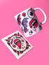 White porcelain 11oz mug and matching 4x4 inch square ceramic coaster set featuring a vibrant, maximalist full-wrap pattern. The design illustrates glossy pink vampire lips with white fangs and red tongues, interspersed with floating eyeballs, purple bats, and small pink skulls against a white background. The mug features a glossy finish and C-handle. The coaster has a matching glazed ceramic top and a brown cork backing. High-contrast pop-art style with pink, purple, and black details.