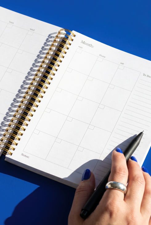 An open spiral-bound notebook with a calendar page is shown, with a hand holding a black pen and a ring visible on the person's finger.