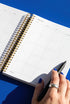 An open spiral-bound notebook with a calendar page is shown, with a hand holding a black pen and a ring visible on the person's finger.