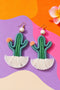 a pair of earrings shaped like cacti, with green and white tassels, set against a colorful background.