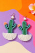a pair of earrings shaped like cacti, with green and white tassels, set against a colorful background.