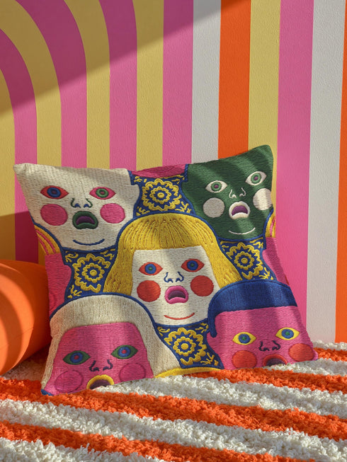 A square decorative pillow cover featuring a faux embroidery pattern of five stylized, expressive doll faces. The central face has yellow hair and pink cheeks. It is surrounded by four other faces in shades of pale beige, green, and bright pink, all with wide staring eyes and open mouths. The design uses intricate, colorful line work to create the illusion of thick woven stitching, but the fabric is a smooth print. Medallions fill the gaps.