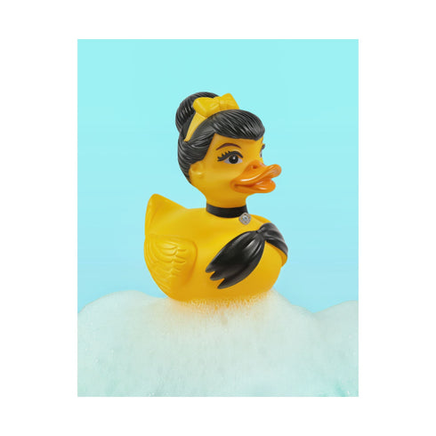 A chic rubber duck styled like Audrey Hepburn’s Holly Golightly, wearing a high bun, yellow bow, black gloves, and a sparkly choker. The duck floats in a pile of foam and is framed in white on a bubbly bathroom shelf with a red tile backdrop and soap dispenser.