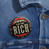 A round pin-back button with a bold, embroidered-style design featuring the phrase "EAT THE RICH" in large, vintage-style lettering. The typography is framed by a set of open red lips with detailed stitching, revealing white teeth. Surrounding the mouth are intricate floral patterns in shades of blue, red, and green, adding a decorative, folk-art-inspired touch. The button has a black background, making the details and lettering stand out