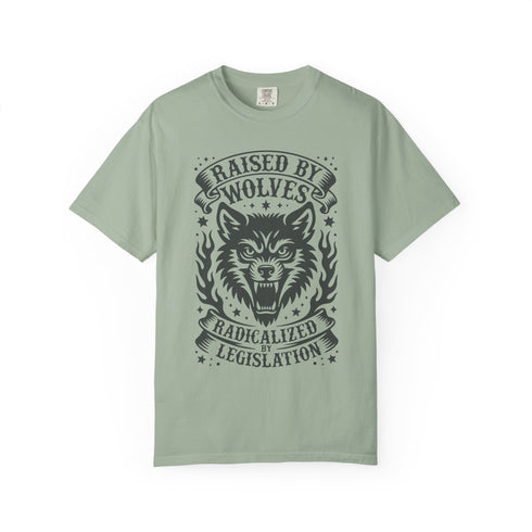 Vintage style graphic t-shirt with a large snarling wolf face printed in dark charcoal ink. A curved banner above reads RAISED BY WOLVES and a banner below reads RADICALIZED BY LEGISLATION, framed by stylized flames and scattered stars. Classic crewneck short sleeve cotton tee with a relaxed fit and seamless sides, available in soft pink, pale green, and ivory. Feminist protest apparel and activist statement tee for reproductive rights, marches, and everyday quiet rage.