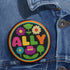 Vibrant ALLY pinback button featuring 70s-style embroidered flowers and lettering in pink, yellow, green, and blue against a black background, surrounded by a rainbow border. A colorful statement accessory combining punk activism and retro flower power aesthetics for backpacks, jackets, and protest wear.