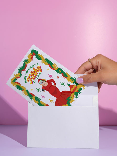 A 5x7 pearlescent holiday card by HoneyDazed. The card illustration, inspired by John Waters, features the drag queen Divine in a red dress with a Santa hat. She points a gun, withtext reads "Have Yourself a Filthy Christmas" in campy font. The card is bordered by garland. Iinside of the card reads: "Season's Sleazings" in red and green text. This kitschy greeting card is a perfect piece of cult film memorabilia and a funny adult humor gift for fans of queer art.