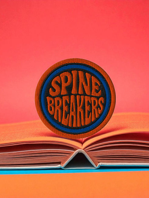 3-inch round embroidered iron-on patch made from a cotton–polyester blend; navy center with blue ring and orange outer ring; stacked orange text reads “SPINE BREAKERS”; merrowed border. Book club patch for paperback addicts, library tote flair, and literary merch collectors; makes a great gift for readers who annotate, dog-ear, and devour novels.