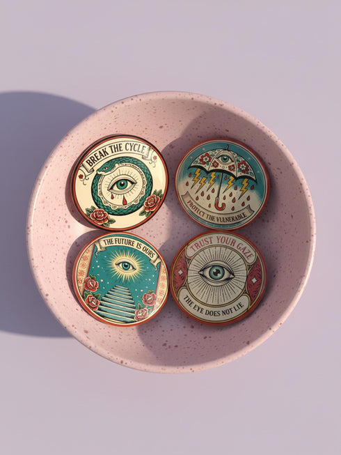 Four 2.25-inch circular metal pinback buttons on a pink textured background. The designs feature vintage tattoo-style art in rust, ochre, gold, and teal with heavy black linework. Designs include a snake Ouroboros "BREAK THE CYCLE," an umbrella eye "PROTECT THE VULNERABLE," a staircase "THE FUTURE IS OURS," and a radial eye "TRUST YOUR GAZE." The pins have a high-gloss finish. Perfect accessory for backpacks, jackets, and lanyards.