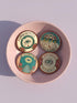 Four 2.25-inch circular metal pinback buttons on a pink textured background. The designs feature vintage tattoo-style art in rust, ochre, gold, and teal with heavy black linework. Designs include a snake Ouroboros "BREAK THE CYCLE," an umbrella eye "PROTECT THE VULNERABLE," a staircase "THE FUTURE IS OURS," and a radial eye "TRUST YOUR GAZE." The pins have a high-gloss finish. Perfect accessory for backpacks, jackets, and lanyards.