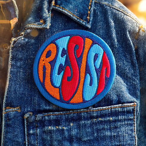 Round embroidered iron-on patch featuring "RESIST" text in bold flame design. Blue border surrounds navy background with orange, red, and blue flame-style lettering. 3x3 inch political activist patch with premium twill fabric construction, perfect for jackets, backpacks, and clothing customization. Anti-establishment protest accessory for resistance movements and political activism demonstrations.