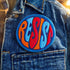 Round embroidered iron-on patch featuring "RESIST" text in bold flame design. Blue border surrounds navy background with orange, red, and blue flame-style lettering. 3x3 inch political activist patch with premium twill fabric construction, perfect for jackets, backpacks, and clothing customization. Anti-establishment protest accessory for resistance movements and political activism demonstrations.