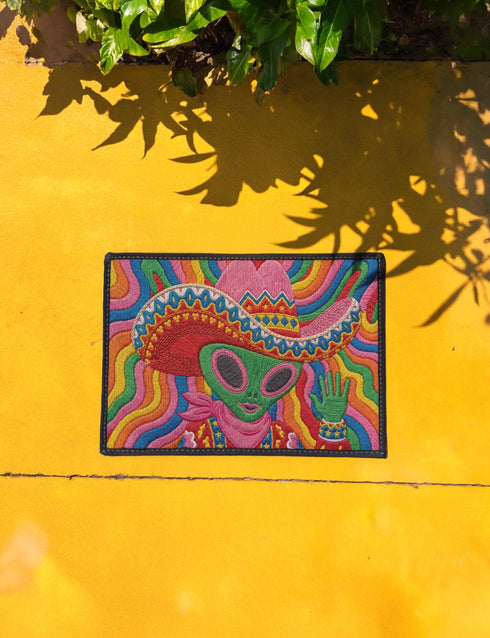 A rectangular doormat featuring a vibrant 2D flat print designed to look like textured yarn embroidery. The central graphic showcases a bright green alien with large black eyes, wearing a pink neck bandana and a highly decorated red, pink, and blue sombrero cowboy hat. The alien has one green hand raised in a friendly wave. The background is a psychedelic pattern of wavy, flowing rainbow stripes in yellow, orange, red, pink, blue, and green. The mat has a sleek black outer edge trim.