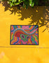 A rectangular doormat featuring a vibrant 2D flat print designed to look like textured yarn embroidery. The central graphic showcases a bright green alien with large black eyes, wearing a pink neck bandana and a highly decorated red, pink, and blue sombrero cowboy hat. The alien has one green hand raised in a friendly wave. The background is a psychedelic pattern of wavy, flowing rainbow stripes in yellow, orange, red, pink, blue, and green. The mat has a sleek black outer edge trim.