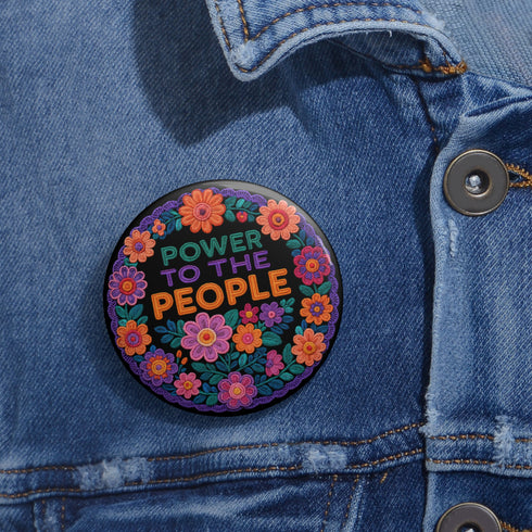 Embroidered-style pinback button featuring the phrase "POWER TO THE PEOPLE" in bold, multicolored satin-stitch letters—green, purple, and orange—framed by a vivid circular floral wreath. Pink, red, and purple flowers with leafy greens radiate around the text. Design has a handcrafted, folk-art feel with a transparent background.