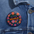 Embroidered-style pinback button featuring the phrase "POWER TO THE PEOPLE" in bold, multicolored satin-stitch letters—green, purple, and orange—framed by a vivid circular floral wreath. Pink, red, and purple flowers with leafy greens radiate around the text. Design has a handcrafted, folk-art feel with a transparent background.