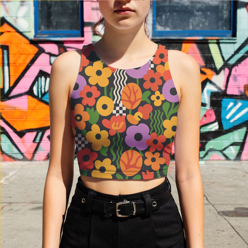 Vibrant retro cropped tank top featuring psychedelic 70s floral pattern with orange yellow purple flowers, black and white checkerboard accents, and wavy green lines on dark background. Body-hugging racerback design made from performance stretch fabric with moisture-wicking properties and UV protection. Perfect for festivals, raves, summer concerts, or bold streetwear styling.