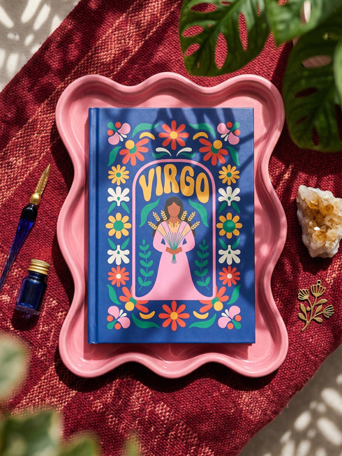 Royal blue hardcover journal featuring a retro folk art design. A center pink arch frames the word VIRGO in golden groovy rounded lettering. Below sits an illustrated maiden with brown skin and dark hair, wearing a pink dress and holding a bundle of golden wheat. A dense floral border wraps the cover with daisies and blooms in orange, marigold yellow, cream white, and pink, accented by green leaves and stems. Matte finish cover with a blue spine reading Virgo. Lined pages inside.