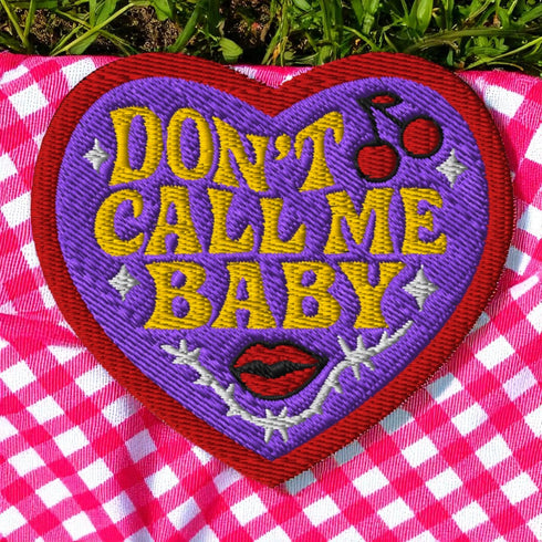 A heart-shaped embroidered patch with a thick red border and a vibrant purple background. In the center, the phrase "DON’T CALL ME BABY" is stitched in a bold, golden-yellow block font. The patch is decorated with small white sparkles, a pair of red cherries with a black musical note, a pair of red lips, and a grey thorny vine design below the text. The overall design has a rebellious, retro, punk aesthetic with a handmade, textured look.