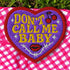 A heart-shaped embroidered patch with a thick red border and a vibrant purple background. In the center, the phrase "DON’T CALL ME BABY" is stitched in a bold, golden-yellow block font. The patch is decorated with small white sparkles, a pair of red cherries with a black musical note, a pair of red lips, and a grey thorny vine design below the text. The overall design has a rebellious, retro, punk aesthetic with a handmade, textured look.