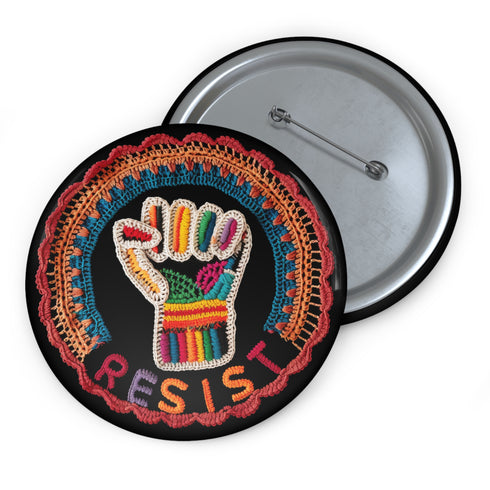 Round pinback metal button with an embroidered-style design on a black background. The center features a raised fist symbol stitched in a multicolored pattern, incorporating rainbow hues and folk-inspired textures. The fist is encircled by a vibrant, lace-like border in shades of blue, orange, and red, resembling intricate embroidery. Below the fist, the word "RESIST" is spelled out in bold, stitched-textured letters in red and orange. The button has a silver metal back with a secure pin closure.