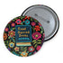 Round pinback button featuring a teal book with "Read Banned Books" in gold lettering on black background, surrounded by vibrant embroidery-style flowers in pink, orange, and blue. Literary protest pin with glossy finish and safety pin backing, available in 2" and 3" sizes for bookish fashion statements.