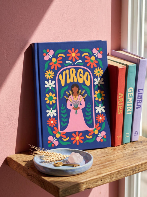 Royal blue hardcover journal featuring a retro folk art design. A center pink arch frames the word VIRGO in golden groovy rounded lettering. Below sits an illustrated maiden with brown skin and dark hair, wearing a pink dress and holding a bundle of golden wheat. A dense floral border wraps the cover with daisies and blooms in orange, marigold yellow, cream white, and pink, accented by green leaves and stems. Matte finish cover with a blue spine reading Virgo. Lined pages inside.