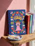 Royal blue hardcover journal featuring a retro folk art design. A center pink arch frames the word VIRGO in golden groovy rounded lettering. Below sits an illustrated maiden with brown skin and dark hair, wearing a pink dress and holding a bundle of golden wheat. A dense floral border wraps the cover with daisies and blooms in orange, marigold yellow, cream white, and pink, accented by green leaves and stems. Matte finish cover with a blue spine reading Virgo. Lined pages inside.