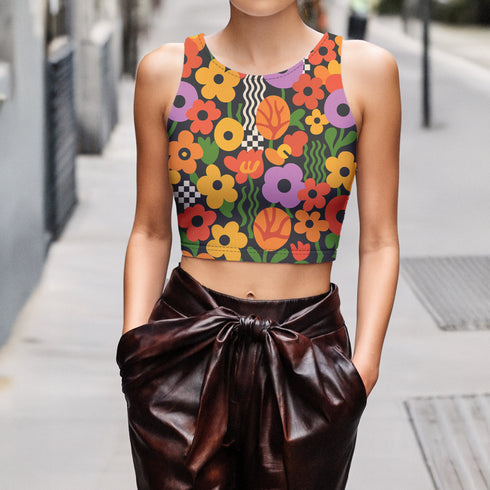 Vibrant retro cropped tank top featuring psychedelic 70s floral pattern with orange yellow purple flowers, black and white checkerboard accents, and wavy green lines on dark background. Body-hugging racerback design made from performance stretch fabric with moisture-wicking properties and UV protection. Perfect for festivals, raves, summer concerts, or bold streetwear styling.