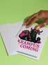 A 5x7 inch Krampus Christmas card shown in two paper finishes. The front features a stylized illustration of the folklore demon Krampus with green skin, black horns, and a pink Santa hat. He carries a large pink sack filled with three skulls over his shoulder. Below him, the words "KRAMPUS IS COMING" are printed in bold pink text. The inside of the card is blank on the left and has the printed text "Hope you've been beautifully bad this year." on the right.