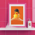 A fine art print of a rubber duck dressed like Alex from A Clockwork Orange, featuring a black bowler hat, blunt bob, and white suspenders. The duck floats in orange-tinged bubbles and is framed in white on a glossy pink tile bathroom shelf.
