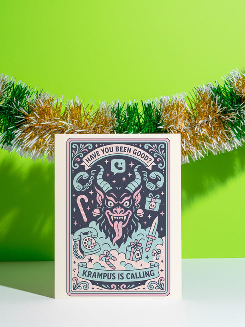 A vertical 5x7 holiday card featuring a retro tattoo style illustration of Krampus in pastel pink, teal, and black ink on shimmery silver pearlescent paper. The central horned demon has a long tongue and is surrounded by vintage rotary phones, daggers, candy canes, and skull gifts. Top text asks Have You Been Good while the bottom banner reads Krampus Is Calling. The interior text reads Let's be honest... Being good is overrated. Merry Christmas.