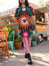 A short-sleeve, above-the-knee t-shirt dress with a straight, structured fit. The dress has a true black background featuring a bold, vibrant graphic pattern. The center displays a large blue and white evil eye surrounded by a bright orange wavy sunburst with a white jagged border. Above and below the eye are bright pink daisy flowers with orange centers, scattered white four-petal stars, and wavy blue and teal green leaf shapes. The fabric is thick, smooth, and holds its clean shape.